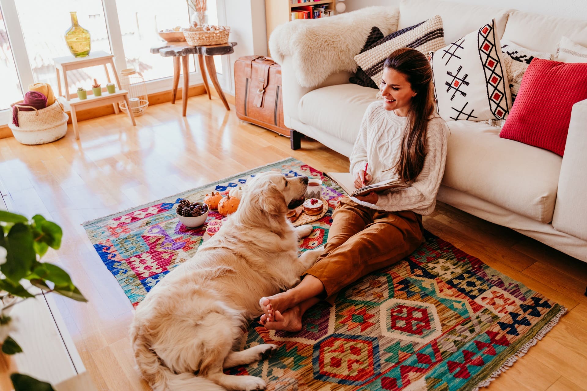 Woman sitting on a colorful area rug with a golden retriever, enjoying snacks and writing in a notebook, surrounded by a cozy living room setting.