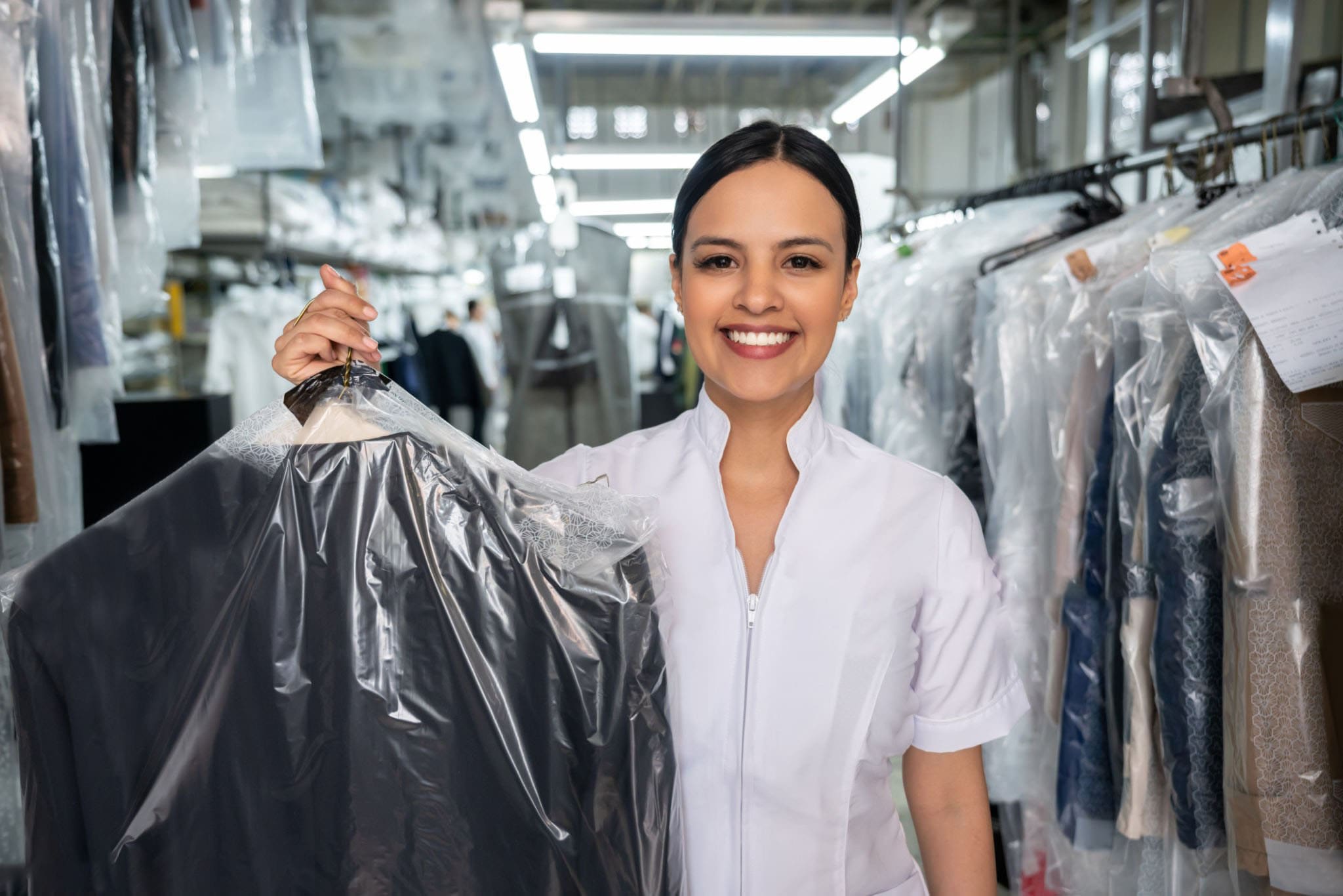 Smiling woman in a uniform holding freshly dry cleaned garment in plastic covering, surrounded by neatly hung clothes in a dry cleaning facility, representing My Best Cleaners' eco-friendly services in Clinton, Maryland.