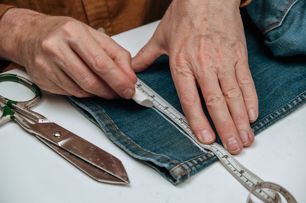 Hands measuring denim fabric with a ruler, scissors nearby, illustrating clothing alteration process for jeans.